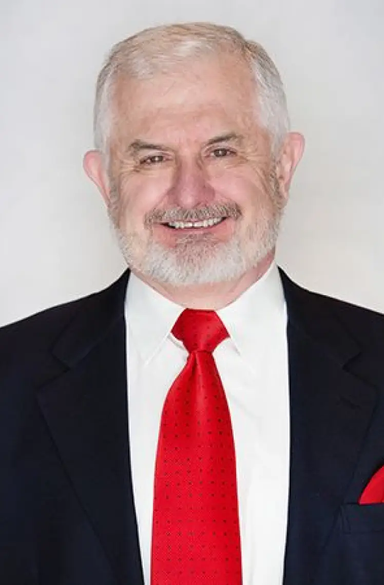 An older man with short white hair and a beard is smiling. He is dressed in a black suit jacket, white shirt, and bright red tie against a plain background, possibly reflecting on the benefits of chronic pain treatment with stem cell injections.