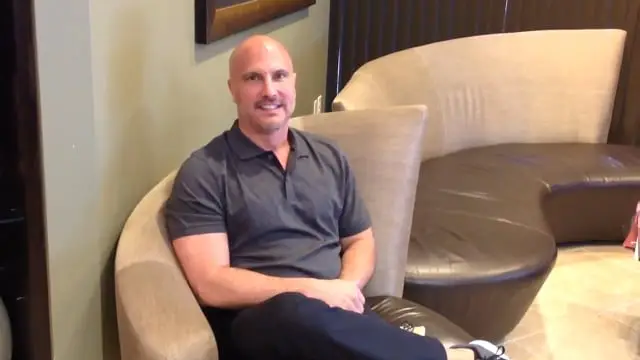 A man with a shaved head and short beard is sitting on a curved beige chair, smiling with his hands folded on his lap. In this modern lounge area, he appears relaxed—perhaps awaiting stem cell therapy Dallas specialists recommend.