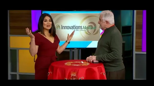 A woman in a red dress gestures while discussing stem cell therapy Dallas with a man in a green sweater. They stand at a round table covered with a red cloth. Behind them, the Innovations Media logo is displayed on a screen.