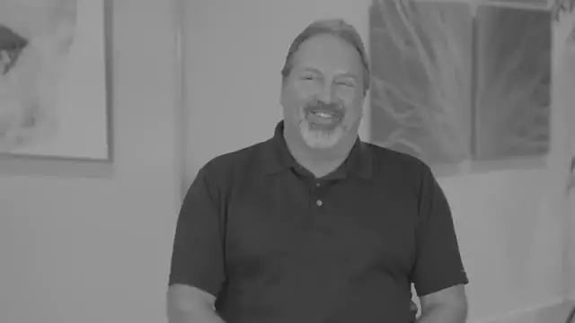 A man with a beard and mustache, wearing a dark short-sleeve polo shirt, sits indoors and smiles at the camera. Abstract artwork is visible on the wall behind him. The image, in black and white, hints at a discussion about stem cell therapy Dallas.