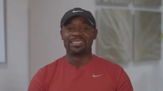 A man wearing a black Nike cap and a red Nike shirt is smiling while sitting indoors, sharing his positive experience with stem cell treatment. The background is softly lit with blurred wall art visible.